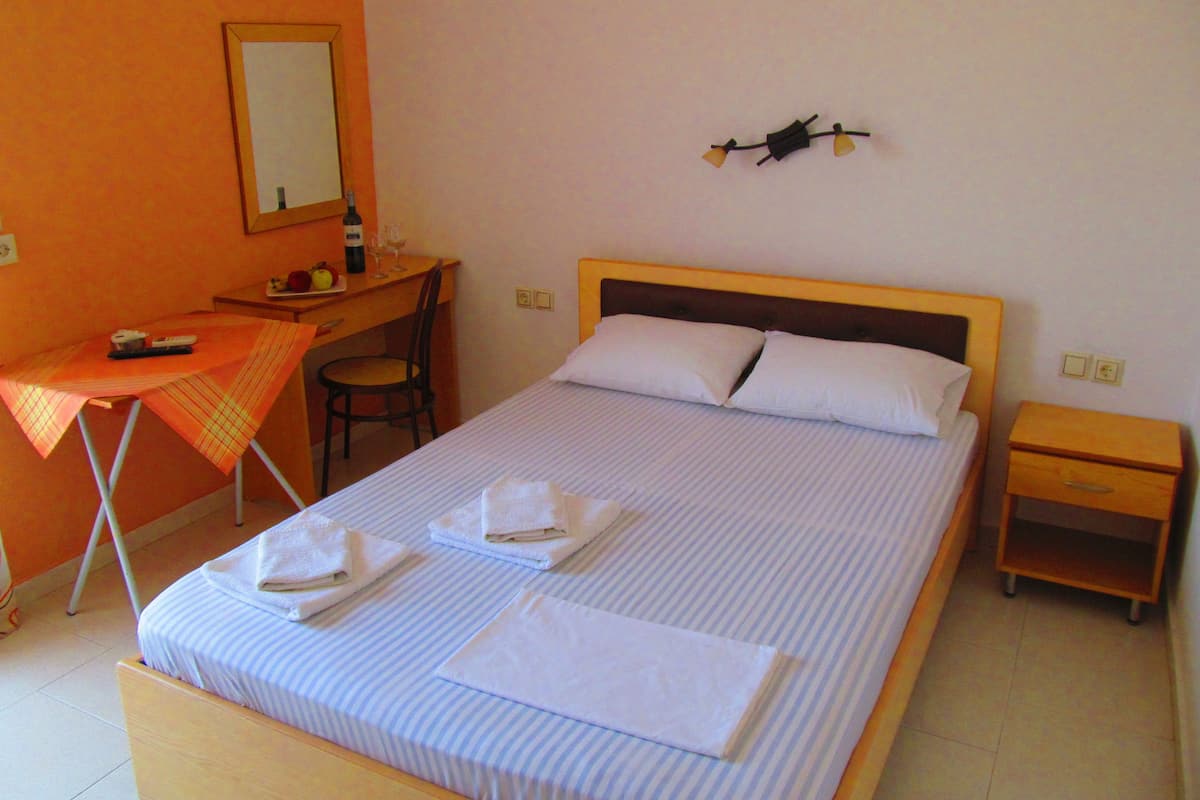 A neatly made bed with pillows and folded towels, a desk with a chair, a small table with a bottle and glasses, and a wall-mounted light fixture.