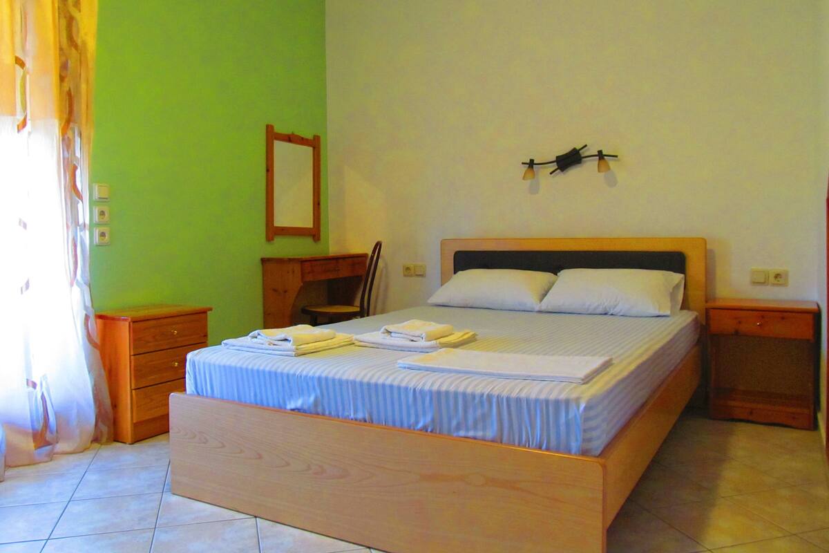 A bedroom with a wooden bed, a nightstand, a chair, a desk, and a mirror on the wall.