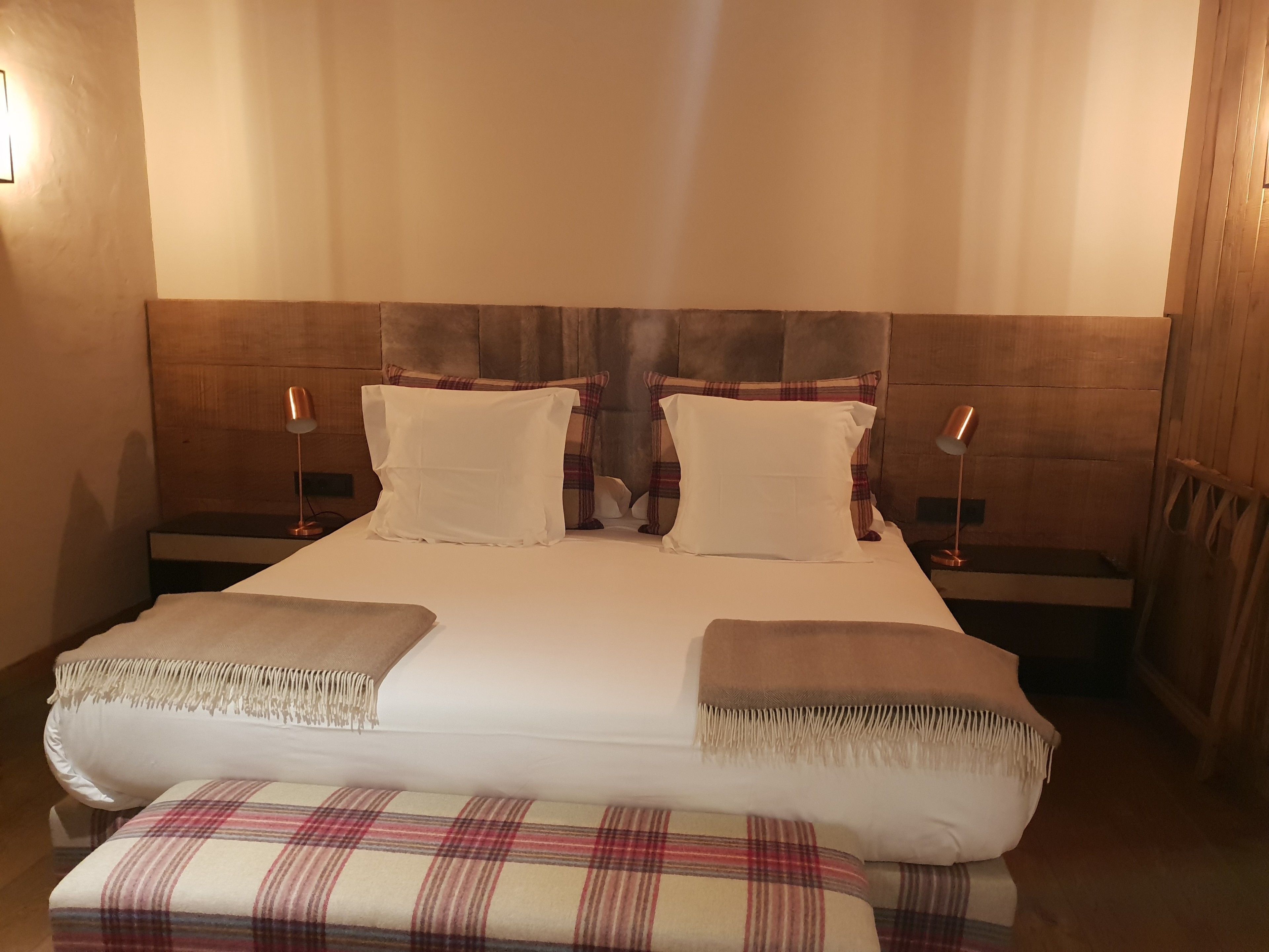 A double bed with two pillows, a bedside table with a lamp, and a bench with a plaid blanket.
