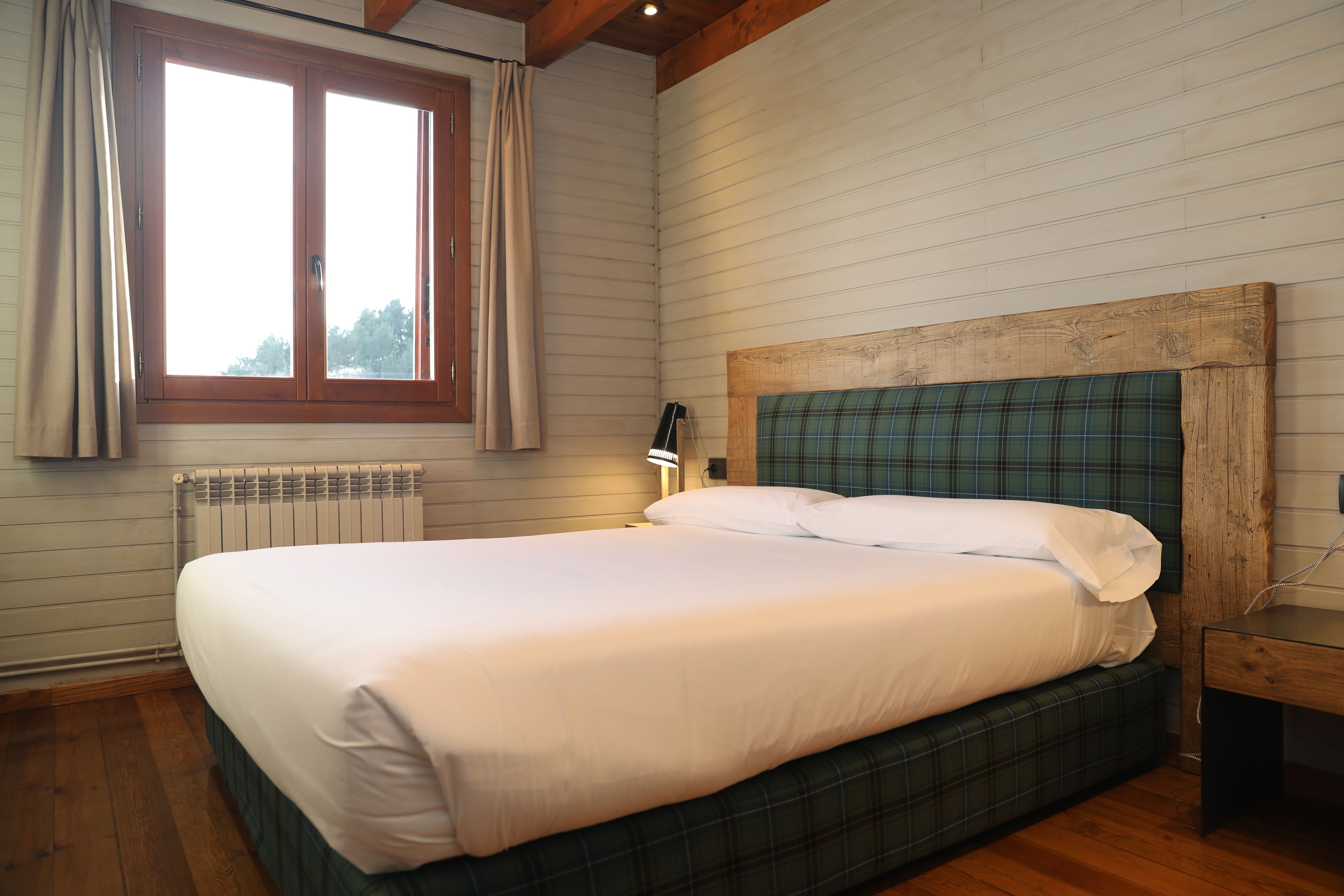 Een net opgemaakt bed met witte lakens in een kamer met houten vloeren en een raam met gordijnen.