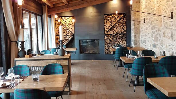 Un restaurant chaleureux avec des poutres en bois, des chaises à carreaux et un mur en pierre. Les tables sont dressées avec des verres et des couverts.
