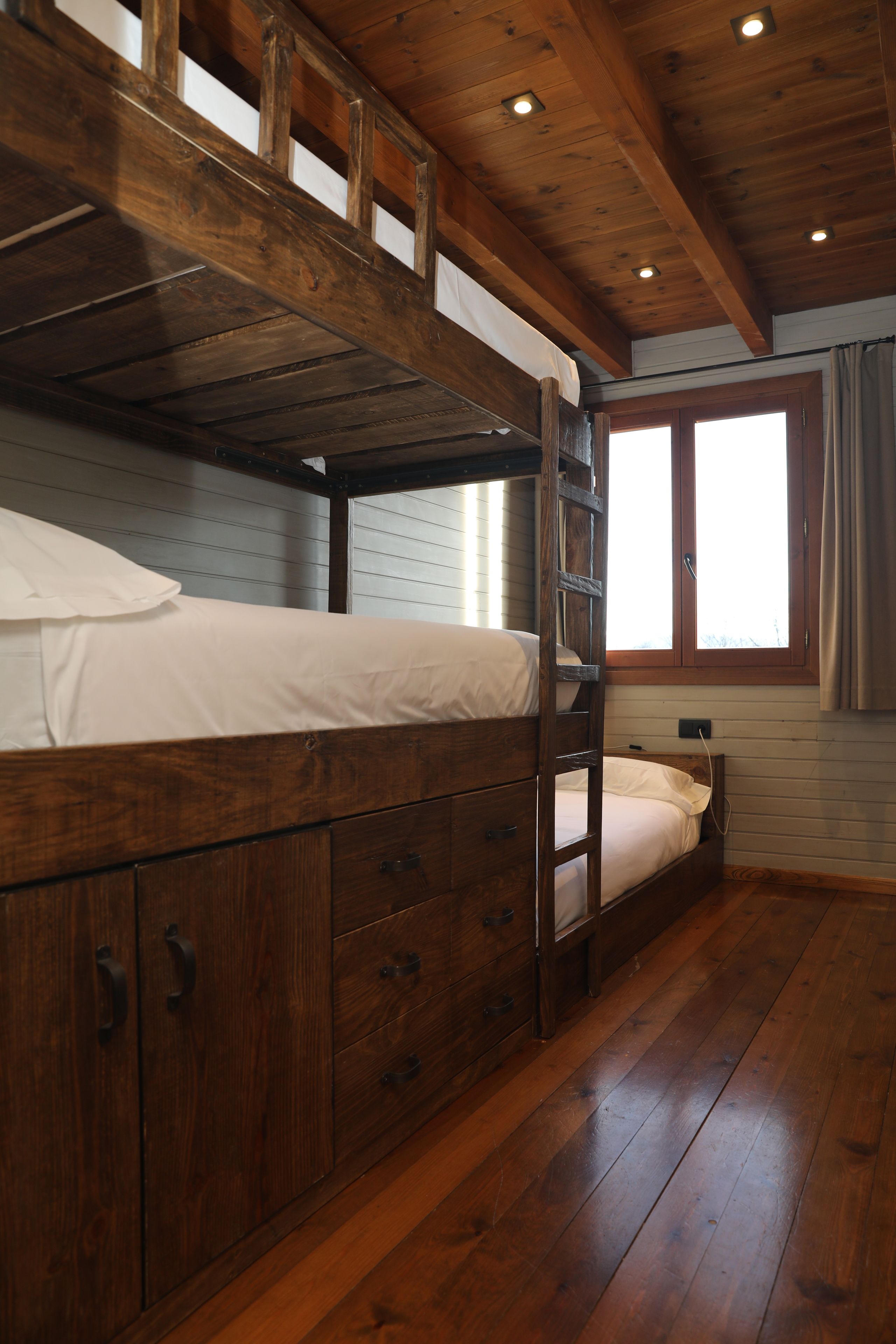 A bunk bed with wooden headboard and drawers, a window with curtains, and a wooden floor.