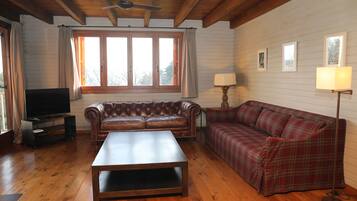 Una sala de estar con techo de madera, un sofá de cuero, un sofá a cuadros, una mesa de centro, un mueble para televisor y una ventana con cortinas.