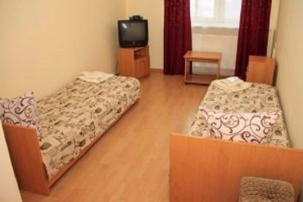 Una habitación pequeña y sencilla con dos camas individuales, una mesita de noche de madera con televisión y una ventana con cortinas.