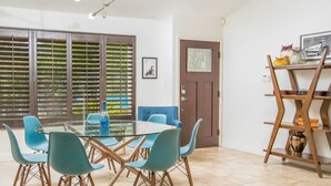 A dining area with a glass table, blue chairs, a brown door, and a wooden shelf with decorative items. - Sunny Palms at Warm Sands - Designer Pool Home (Palm Springs)
