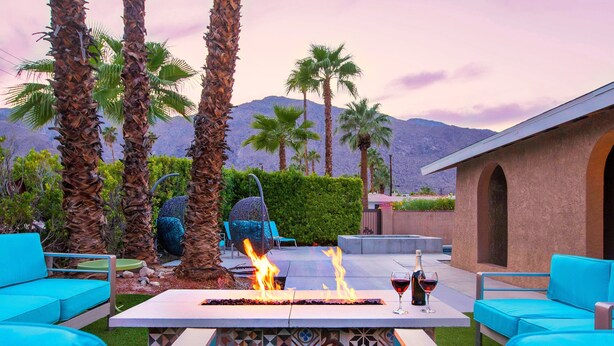 A patio with a fire feature, two glasses of red wine, and blue outdoor furniture. - Sunny Palms at Warm Sands - Designer Pool Home (Palm Springs)