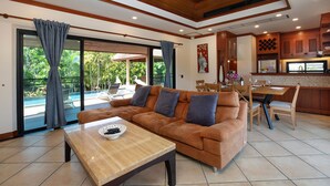 A spacious living area with a sofa, a coffee table, and a dining table with chairs. There is a kitchen area with wooden cabinets and a large window overlooking a pool. - Boutique 3BR Pool Villa Bianca NaiHarn (Rawai)