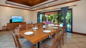 A dining area with a wooden table set for four, a television on a wooden cabinet, and a view of the outdoors through large windows. - Boutique 3BR Pool Villa Bianca NaiHarn (Rawai)