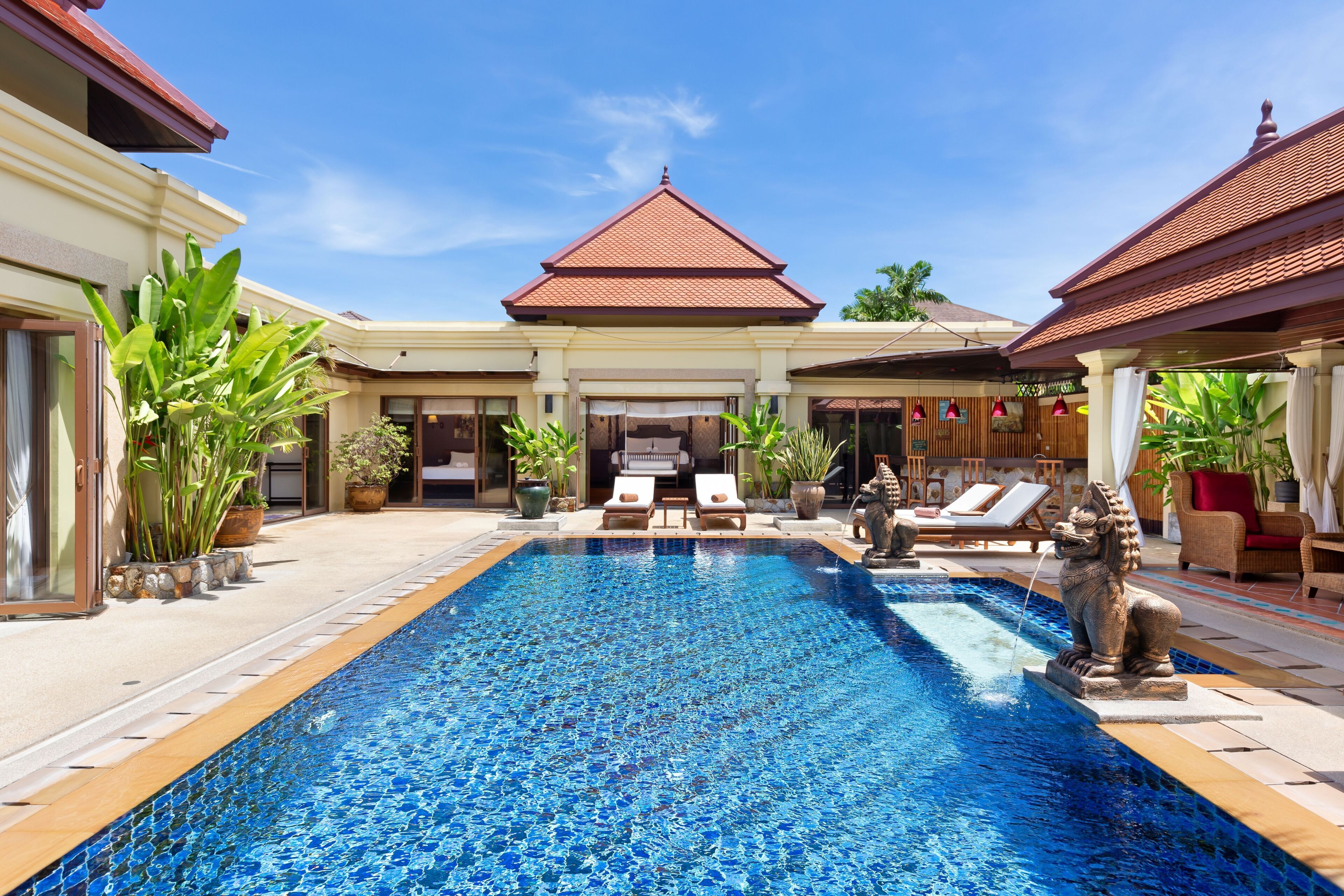 A swimming pool with lounge chairs and a statue, surrounded by potted plants and a building with a tiled roof.