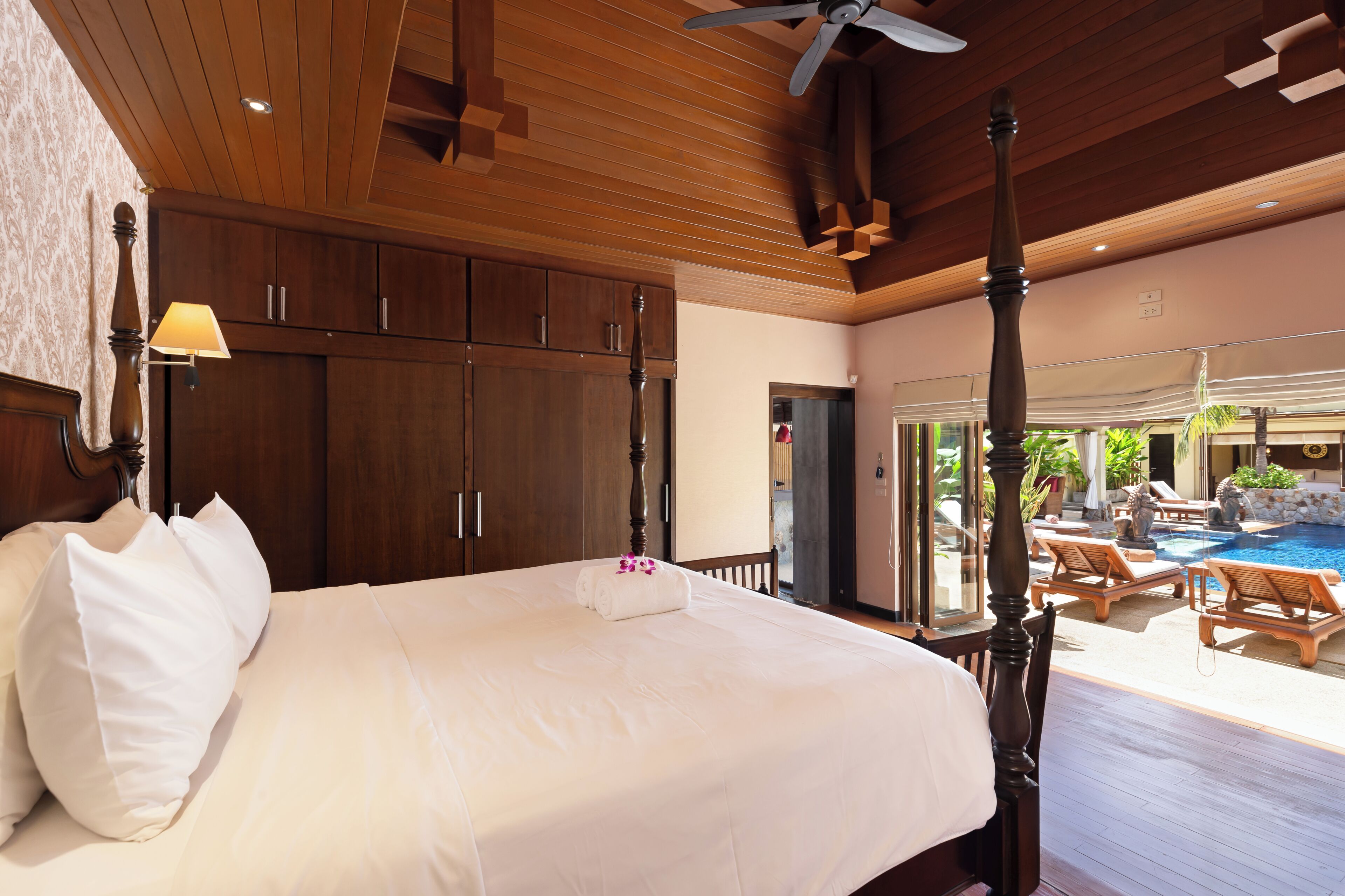 A four-poster bed with a canopy, wooden ceiling, and a view of a pool area.