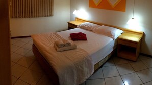 A hotel room with a bed, two bedside tables, a nightstand, a window with blinds, and a wall-mounted artwork. - Residencial Cachoeira Flats (Florianópolis)