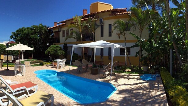 A pool area with lounge chairs, a covered seating area, and a building with a yellow facade. - Residencial Cachoeira Flats (Florianópolis)