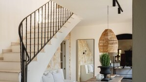 Un escalier avec une rampe en métal noir, un canapé blanc avec des coussins, une table en bois sur laquelle est posée une plante en pot, et une lampe en osier.