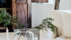 Un salon chaleureux avec une porte en bois, un canapé blanc, un vase en verre contenant des branches, une bougie et un livre posé sur une table.