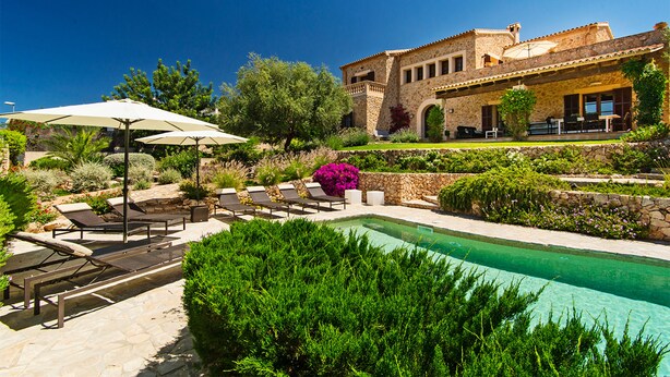 Un espace aménagé au bord de la piscine, avec des chaises longues et des parasols, un bâtiment en pierre doté d’arcades et un jardin bien entretenu.