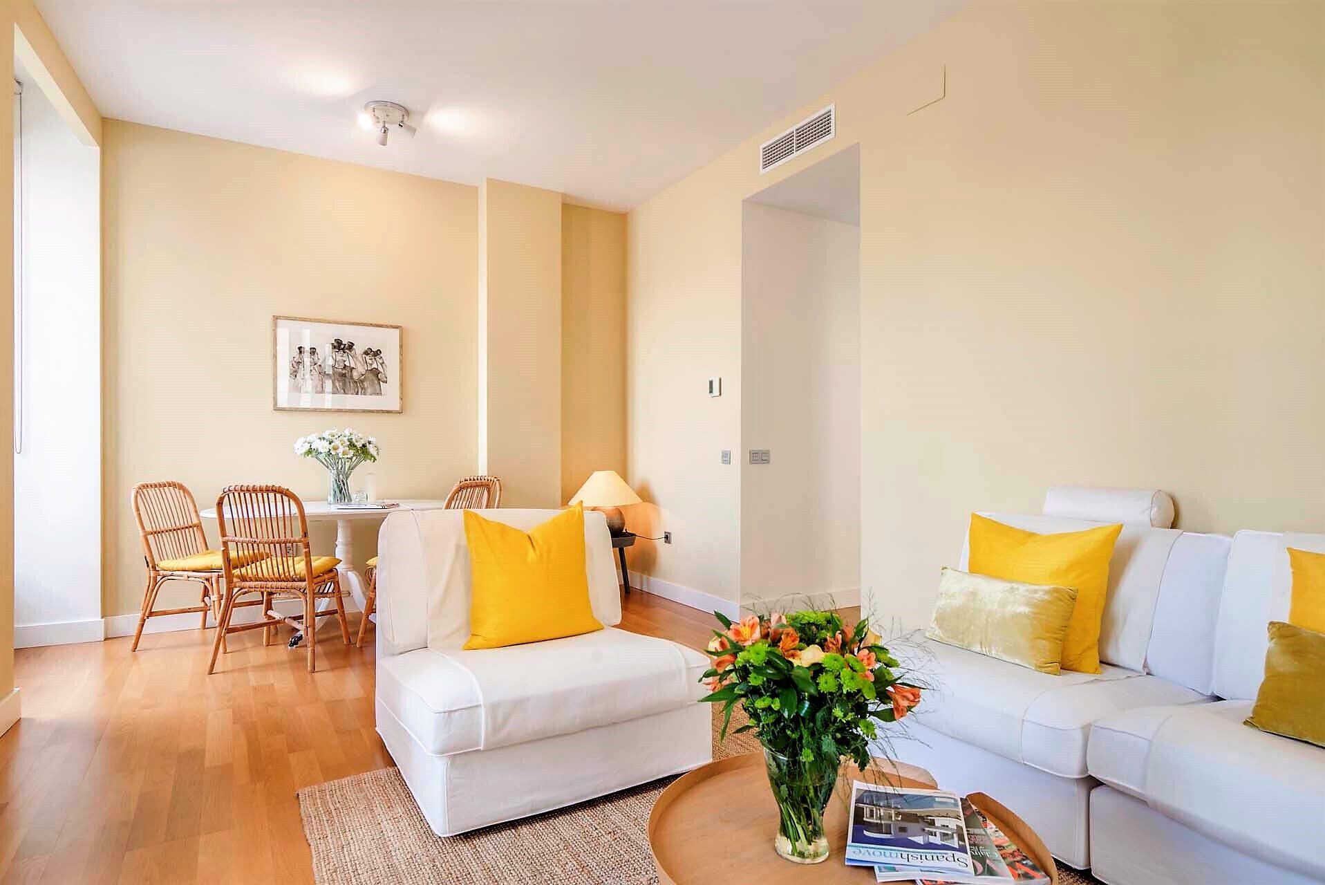 A living room with a white sofa, yellow cushions, a dining table with chairs, and a framed picture on the wall.
