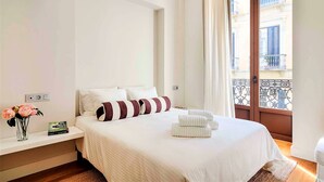 Habitación de hotel con cama, mesita de noche, un florero con flores y vistas a un balcón con edificios.
