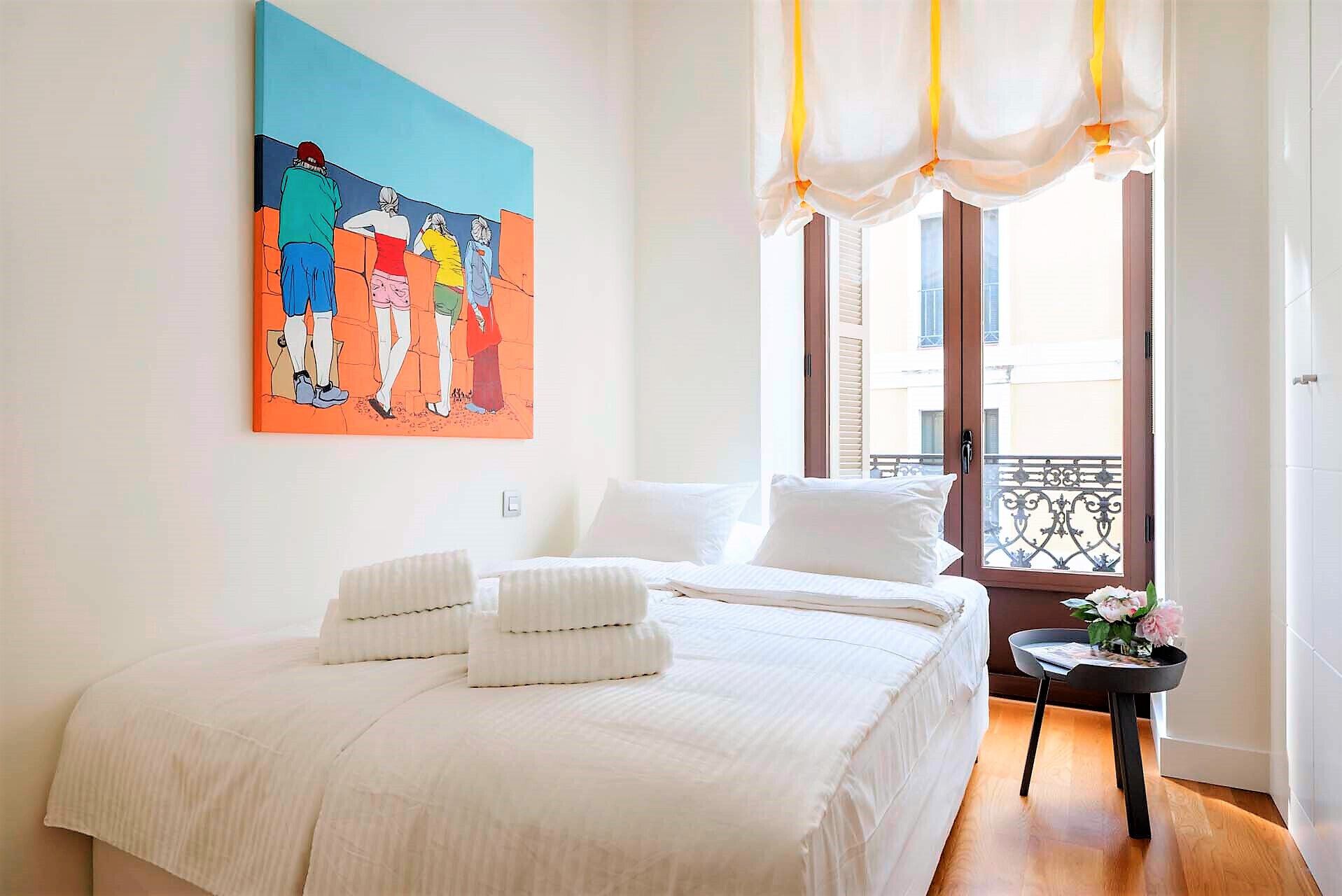 Habitación de hotel con cama, un cuadro, una ventana con cortinas y una mesita con un florero.