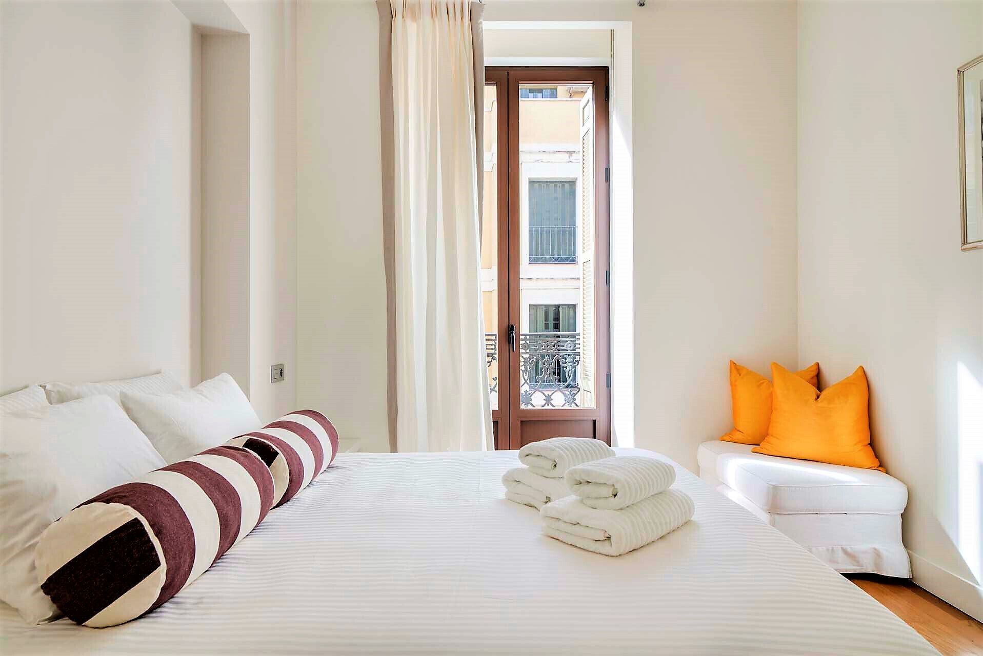 A hotel room with a bed, white curtains, a view of a balcony, and a white sofa with orange cushions.