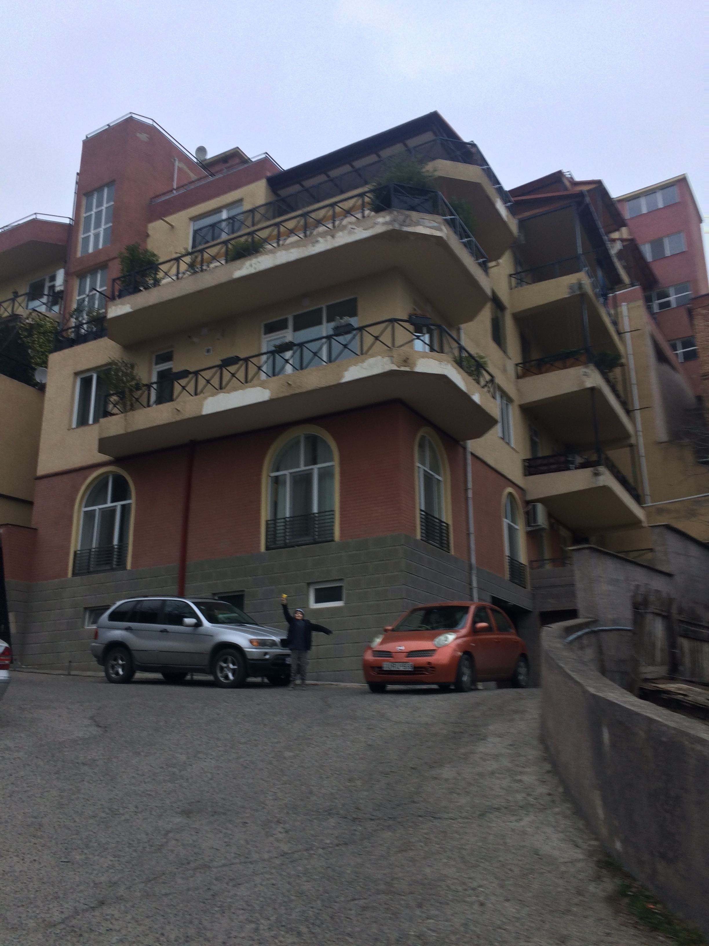 Un immeuble résidentiel à plusieurs étages, doté de balcons, avec des voitures garées et une personne debout près de l’entrée.