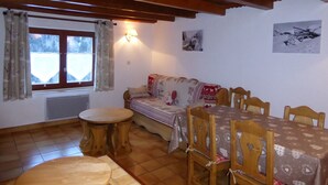 A cozy dining area with a wooden table and chairs, a round wooden table, a sofa with patterned cushions, and a window with curtains.