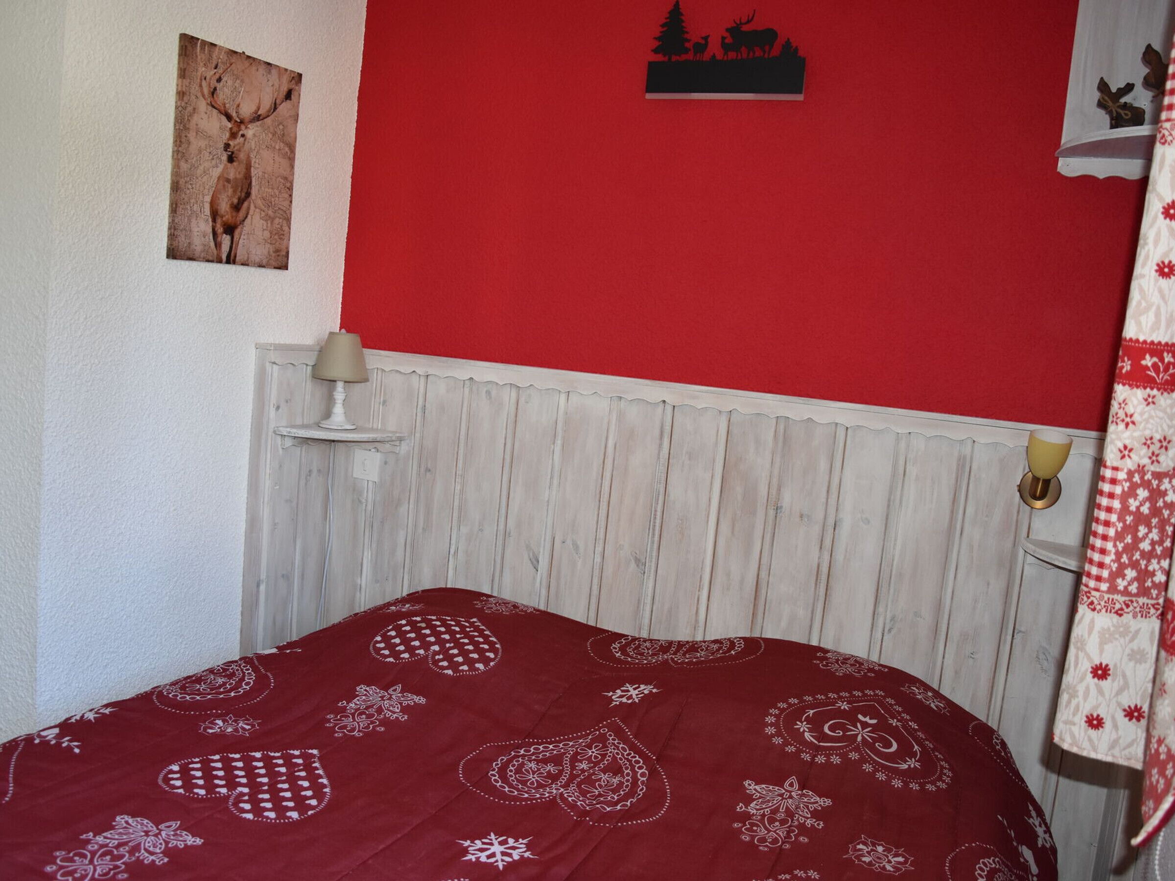 Ein Schlafzimmer mit roter Wand, einem Holz-Kopfteil, einem Bett mit rot gemustertem Überwurf, einem kleinen Tisch mit Lampe sowie einer Wanddekoration mit einem Reh und Bäumen.