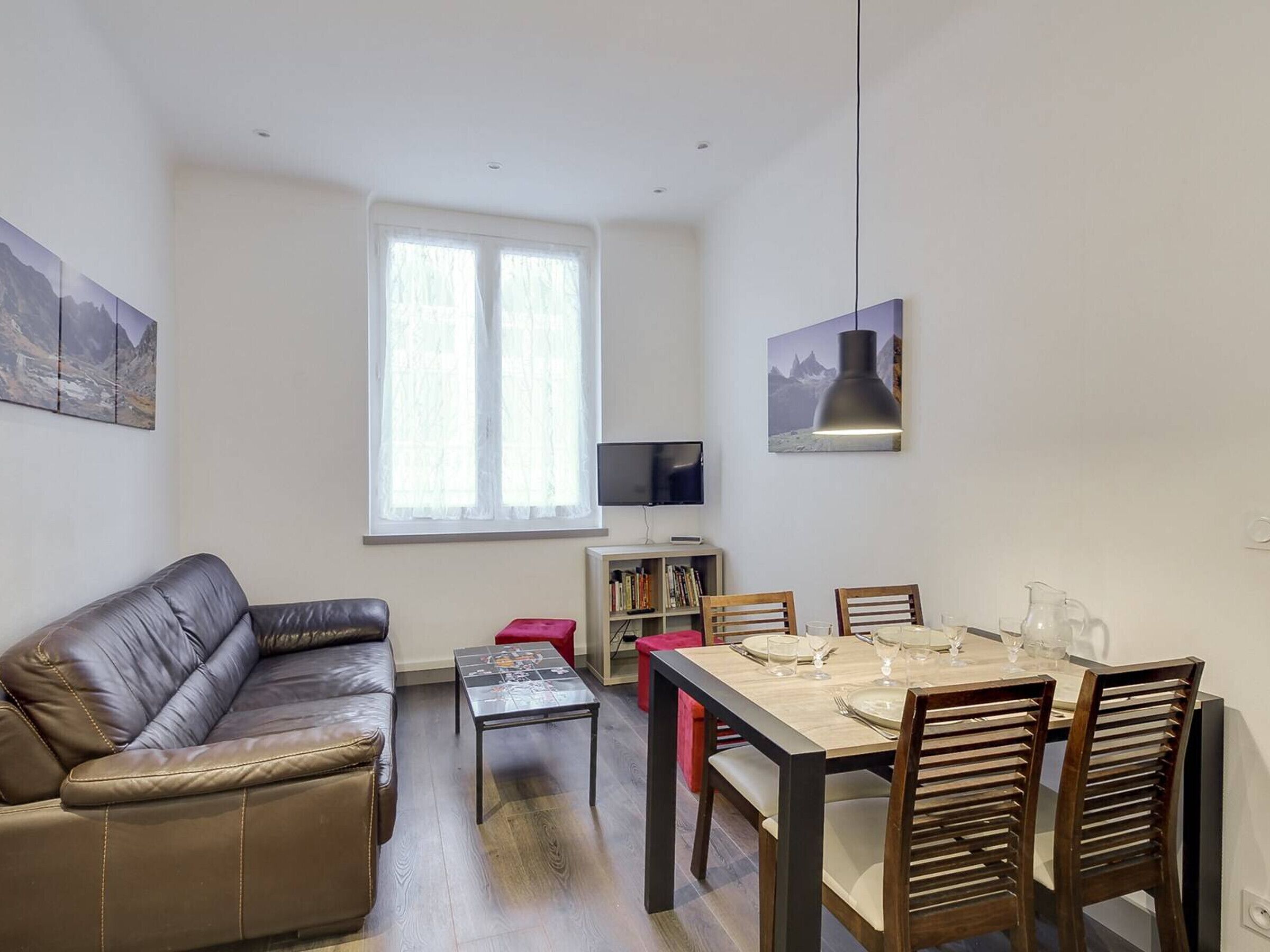 Un salon moderne avec un canapé en cuir, une table à manger pour quatre personnes, une bibliothèque et une grande fenêtre ornée de rideaux légers.