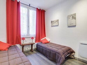 Une chambre avec deux lits, des rideaux rouges, une fenêtre avec des rideaux blancs, une petite table avec des lampes et deux œuvres d’art accrochées au mur.