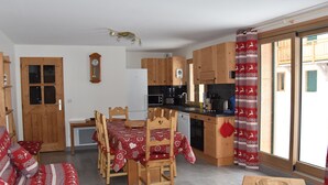 Una sala de estar acogedora con sofá, mesa de comedor y sillas, una cocina con armarios de madera y electrodomésticos, y un amplio ventanal con cortinas.