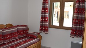 Una habitación con cama de madera, cortinas con estampado rojo y blanco, una ventana con vista a edificios y una rejilla en la pared.
