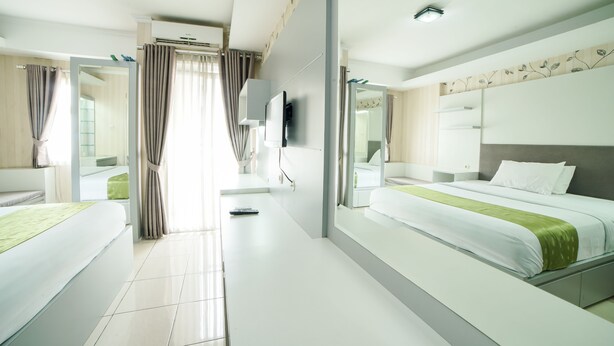 A hotel room with a large bed, a smaller bed, a desk, a mirror, a TV, and a bathroom with a shower and toilet. - Apartment Bogor Valley (Bogor)