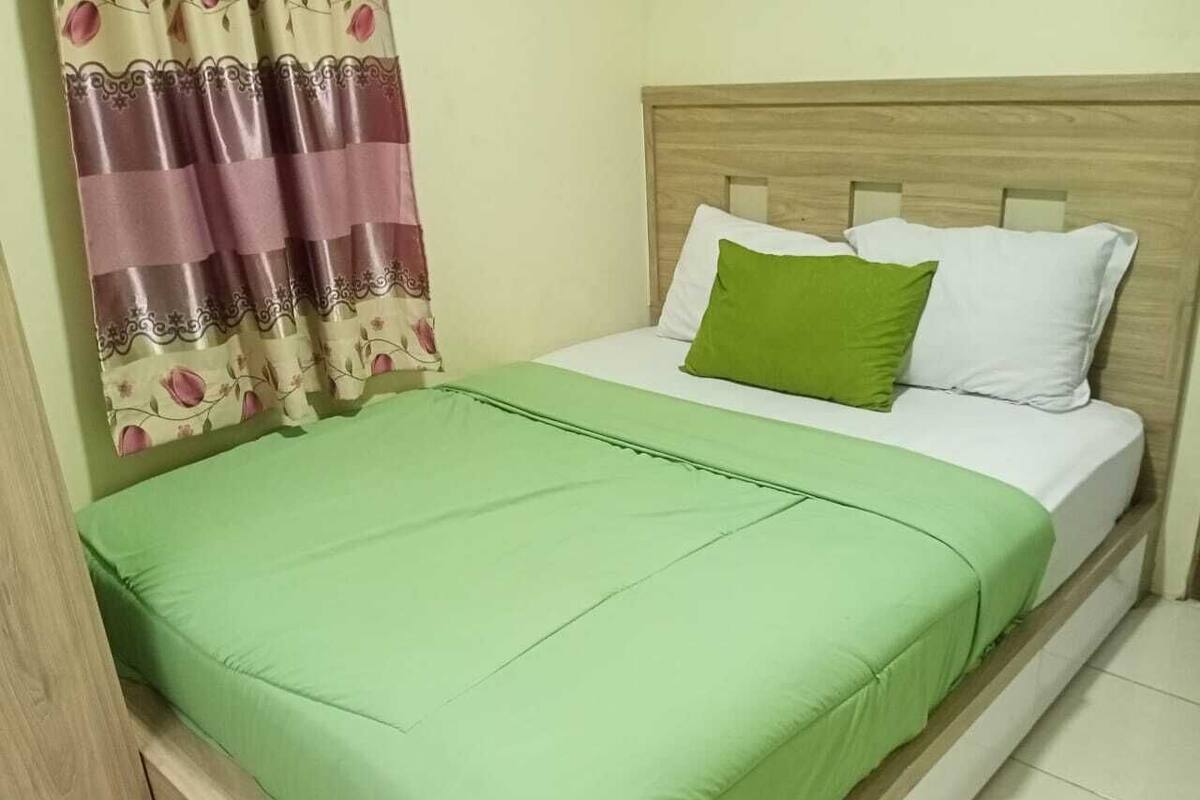 A bed with a green bedspread and pillows, a wooden headboard, and a curtain with a floral pattern.