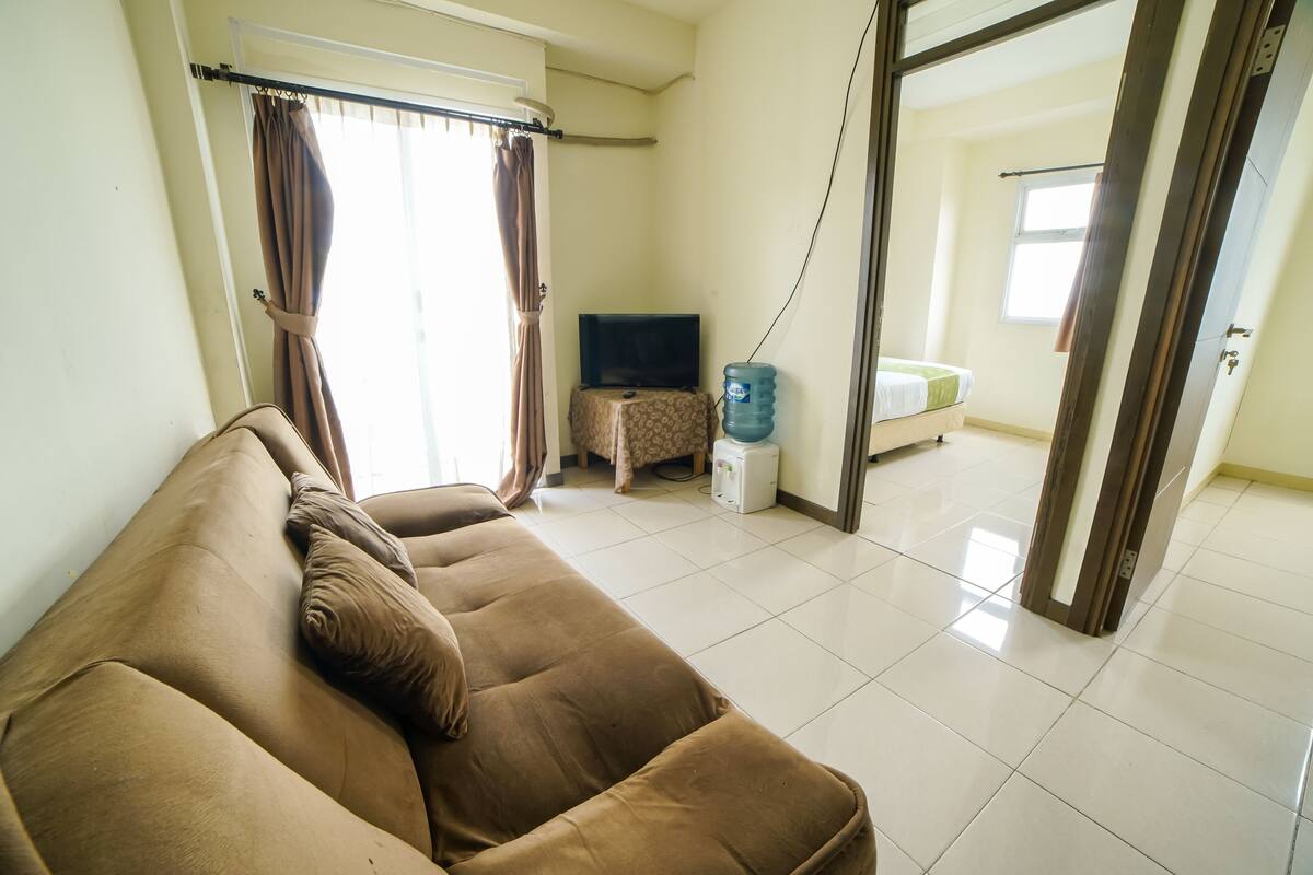 A living room with a brown sofa, a flat-screen TV, a water dispenser, and a view to a bathroom through an open door.