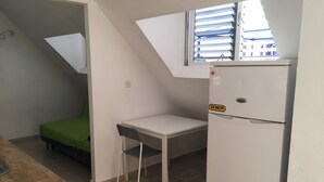 A small kitchenette with a white refrigerator, a green bed, and a white table with a chair.