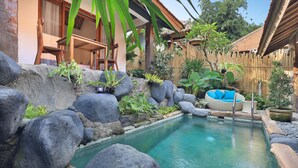 A pool area with large rocks, a wooden deck, and a lounge chair with blue cushions. - Batu Agung Villas (Ubud)