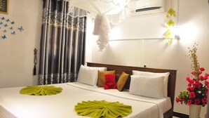 A neatly made bed with white linens and green decorative pillows, a bedside lamp, and a vase of red flowers. - Sigiri Saman Homestay (Sigiriya)
