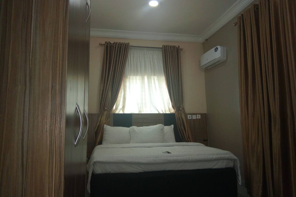 A hotel room with a bed, curtains, an air conditioning unit, and a window with curtains.