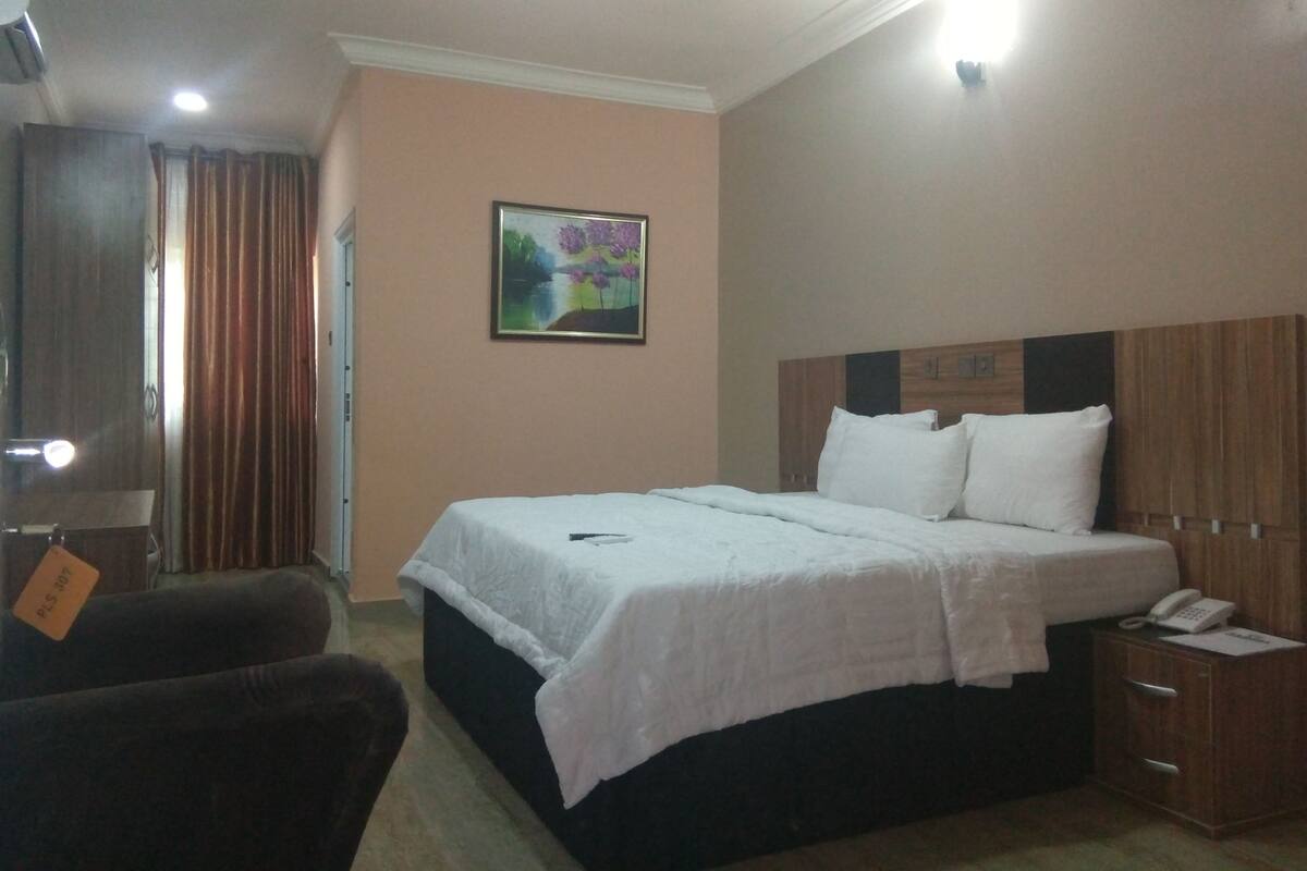 A hotel room with a bed, a nightstand, a chair, a painting on the wall, and a door.