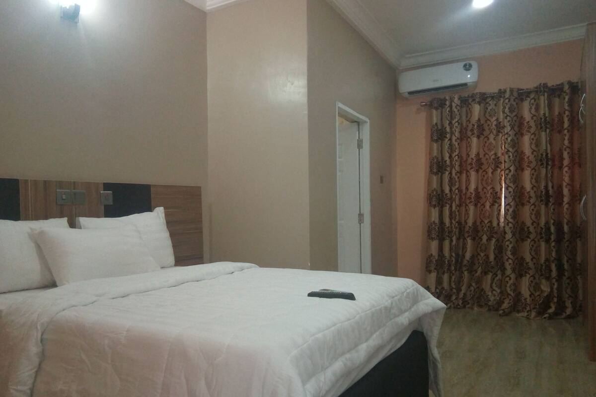 A hotel room with a bed, a remote control on the bed, an air conditioning unit on the wall, and a door.