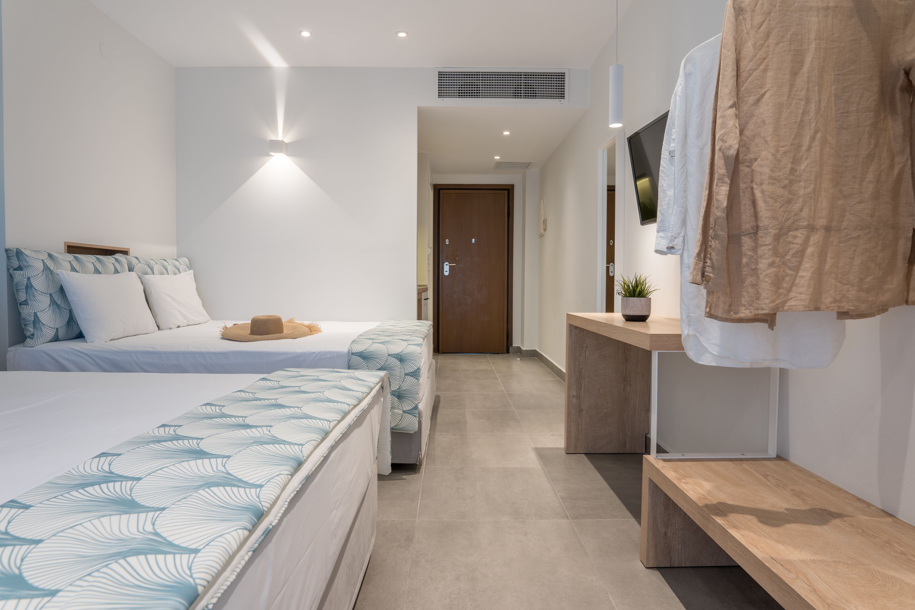 A hotel room with two beds, a wooden bench, a small table, a TV, and a door leading to another room.