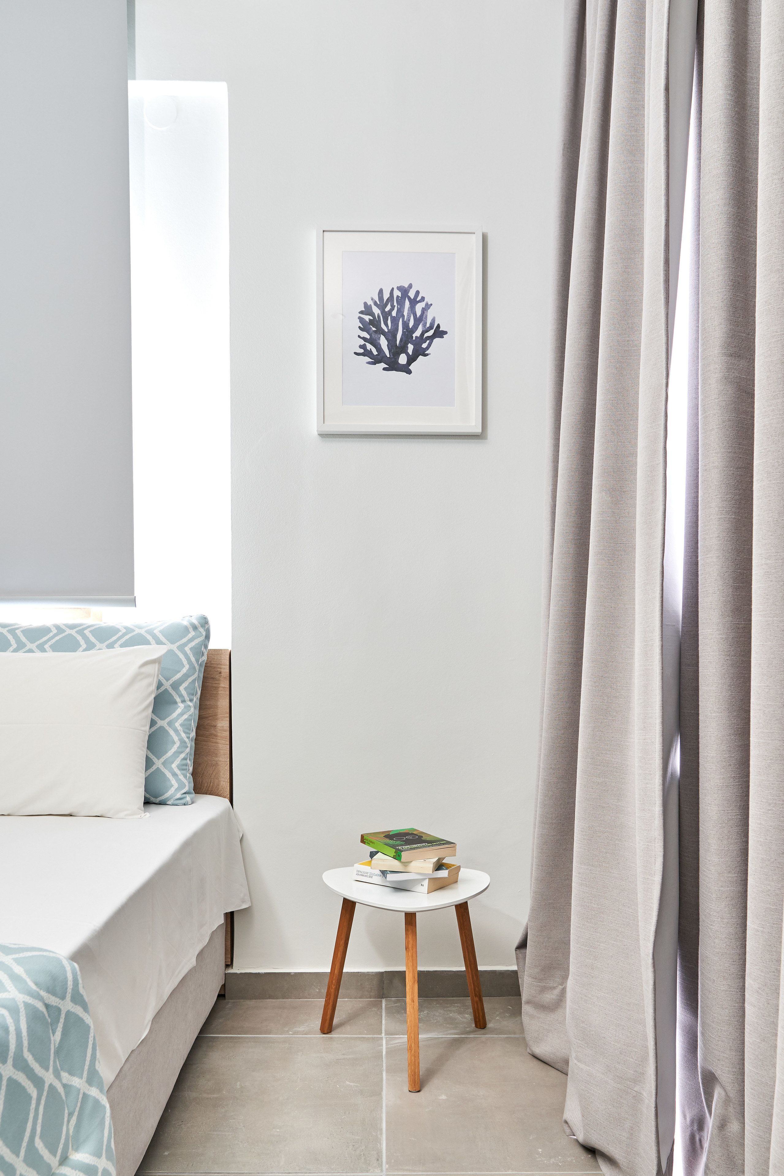 A bedroom with a bed, a small bedside table with books, a framed artwork on the wall, and a window with curtains.