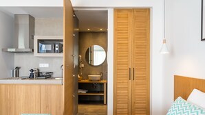 A modern hotel room with a bed, a wooden wardrobe, a bathroom with a sink and mirror, and a kitchenette with a microwave.