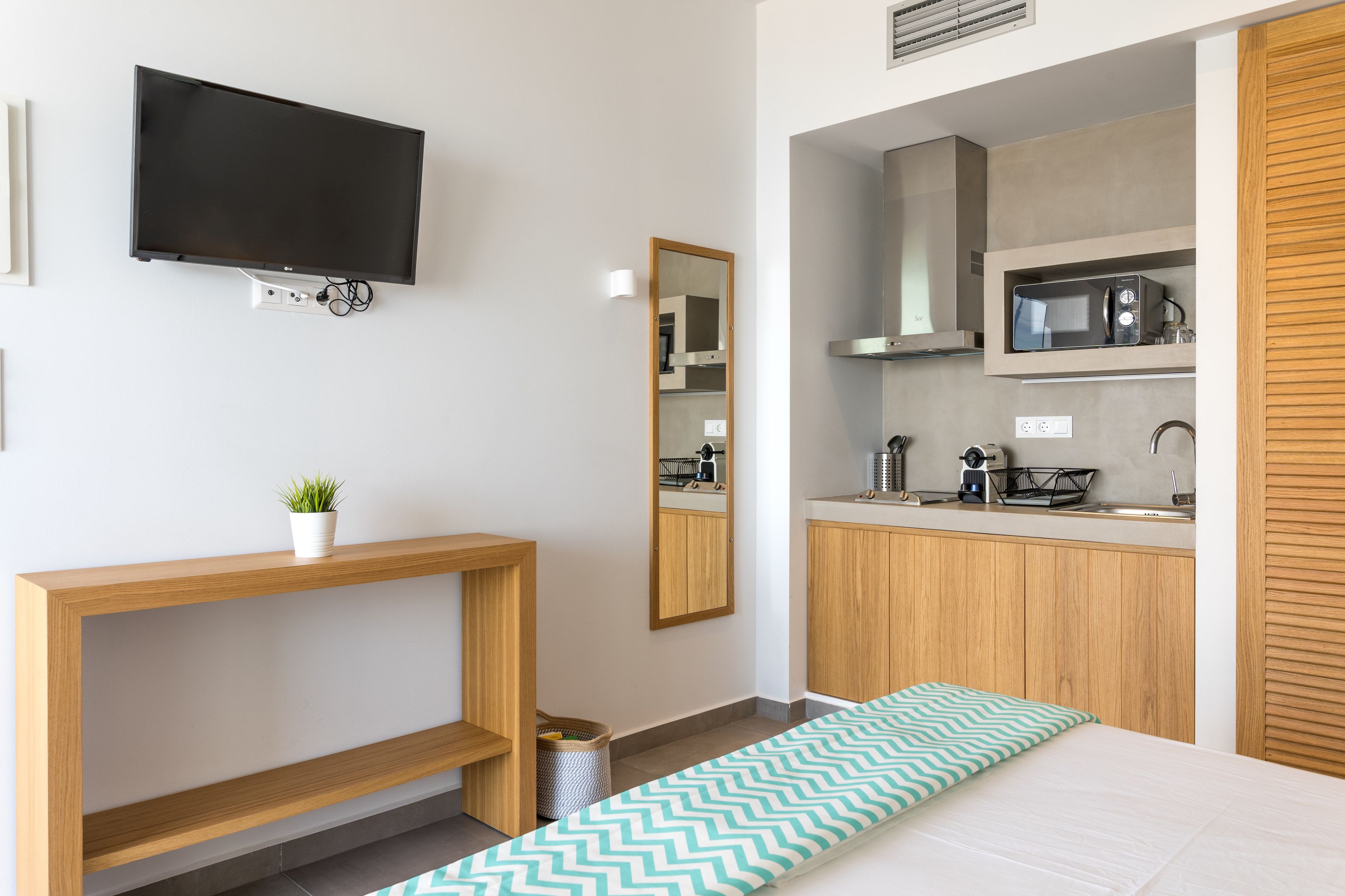 A modern hotel room with a TV mounted on the wall, a wooden console table, a small potted plant, a mirror, a kitchenette with a microwave, and a bed with a patterned bedspread.