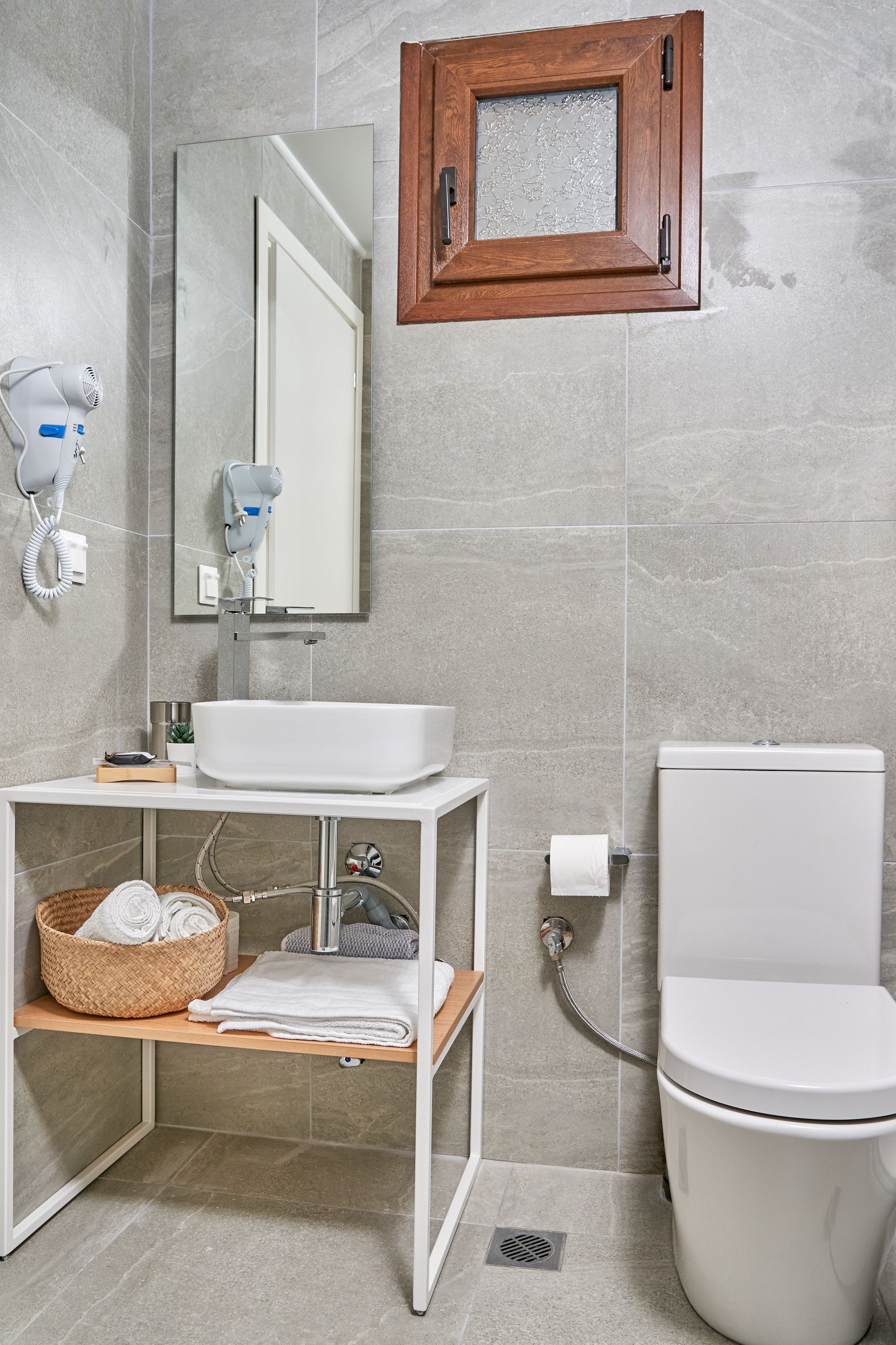 Uma casa de banho moderna com uma pia branca, um espelho, um armário de maquilhagem em madeira, uma sanita e um secador de cabelo na parede.
