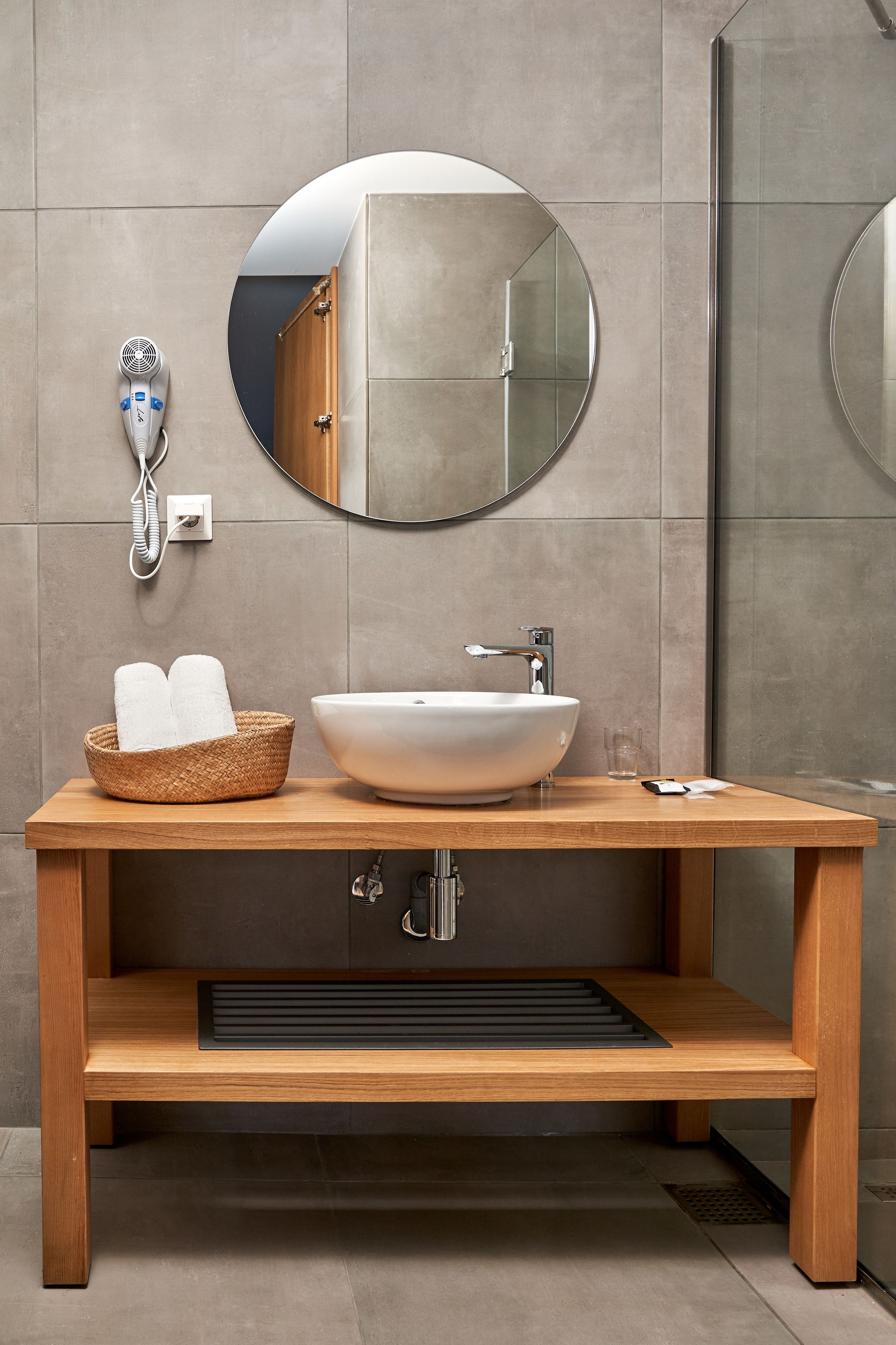 Ein modernes Badezimmer mit einem Holzkosmetikschrank, einem runden Spiegel, einem weißen Waschbecken und einem Fön an der Wand.