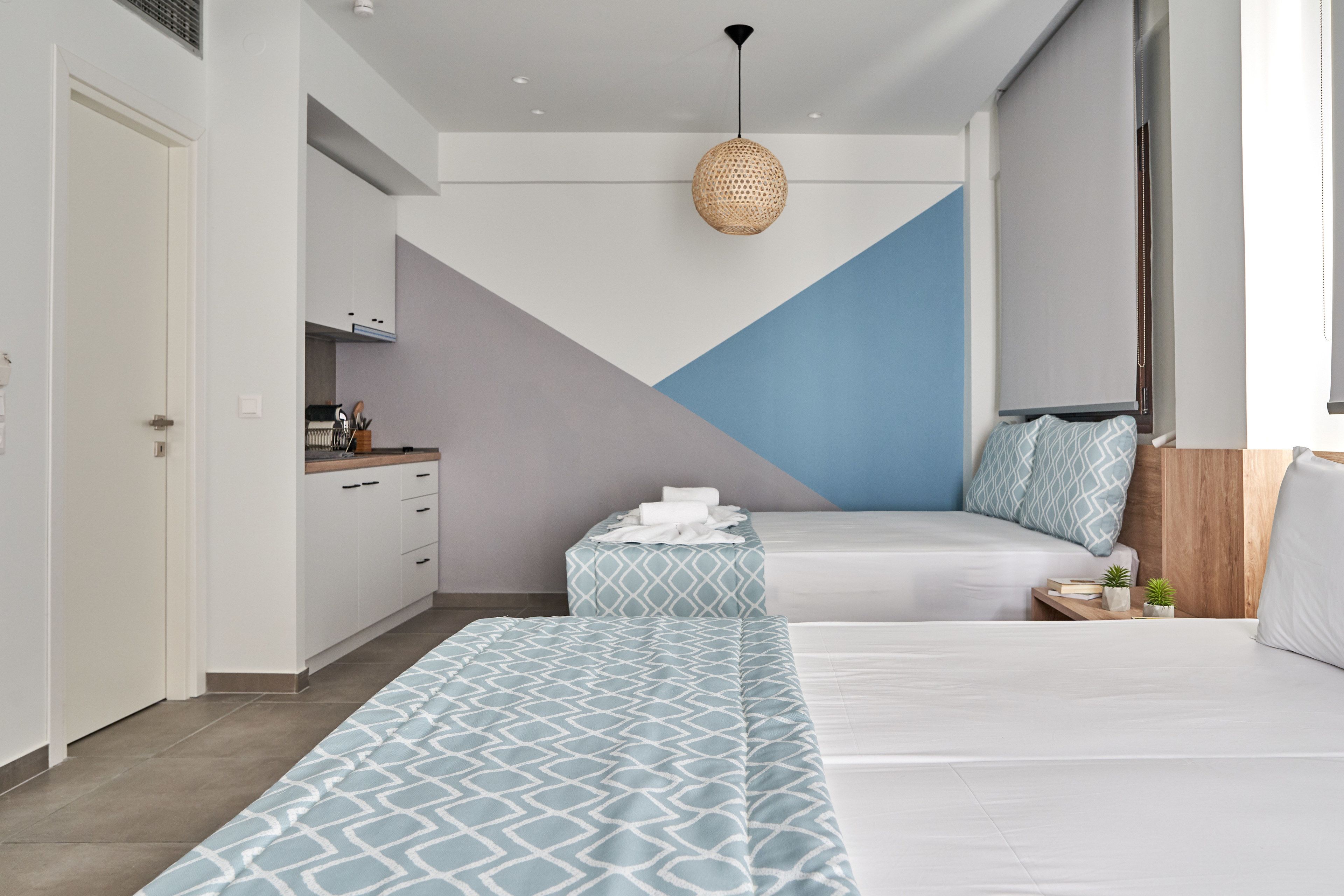 Una habitación de hotel moderna con un diseño geométrico en la pared, una kitchenette, una cama con ropa de cama estampada y un colgante de ratán.