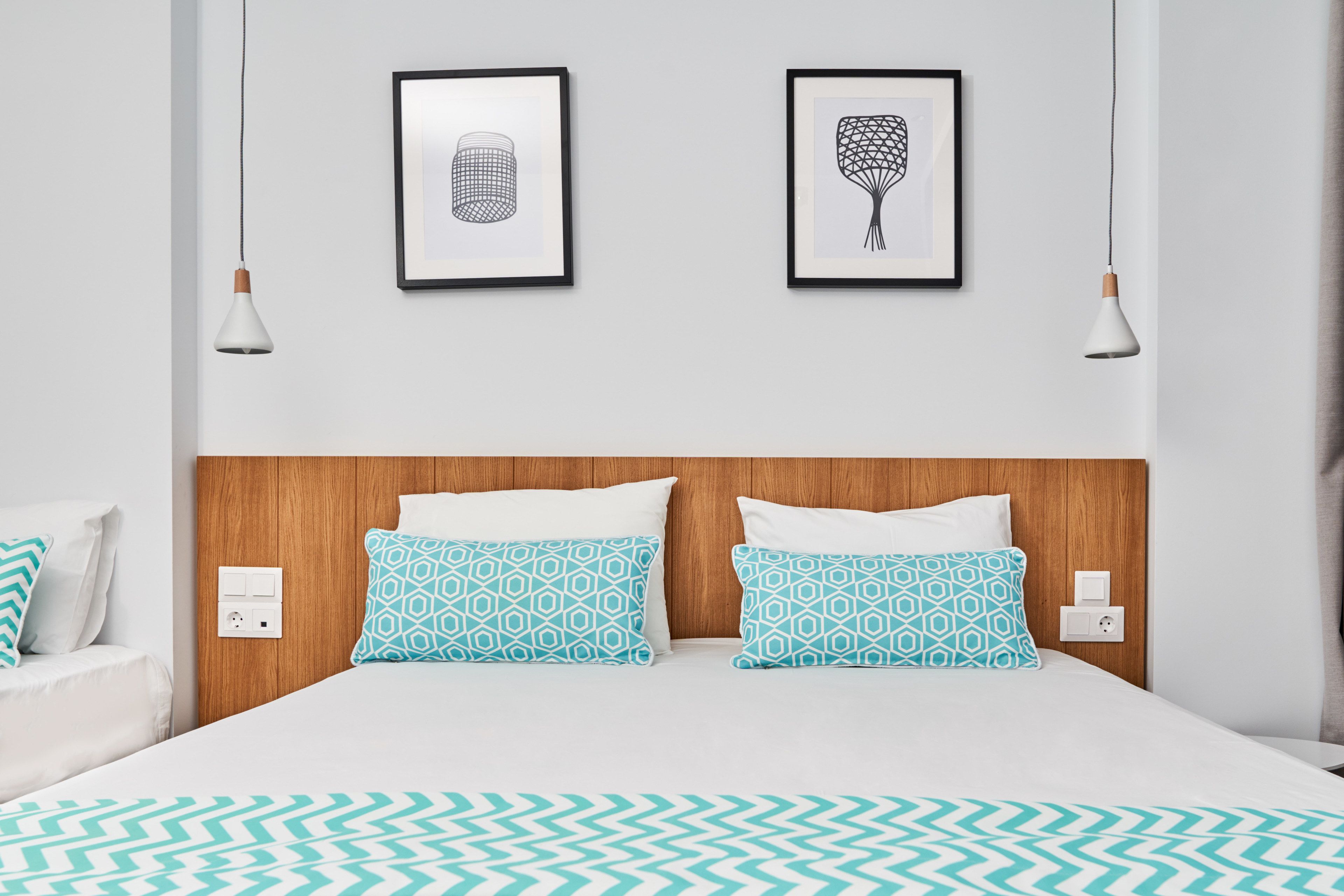 Ein Hotelzimmer mit einem Holzkopfteil, zwei gemusterten Kissen, einer weißen Tagesdecke und zwei gerahmten Kunstwerken an der Wand.