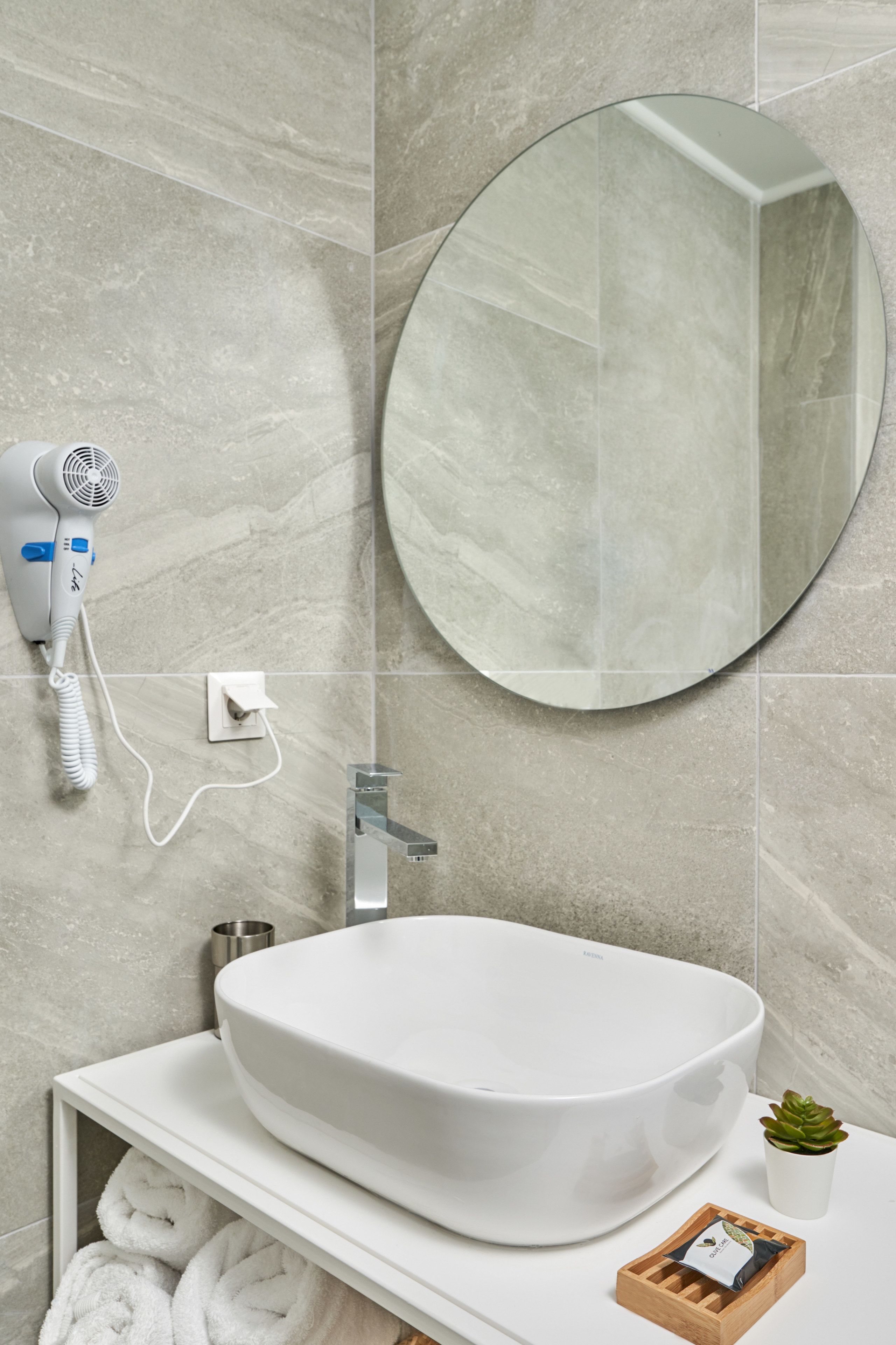 Une salle de bain avec un lavabo blanc, un miroir rond, un sèche-cheveux et une petite plante en pot.