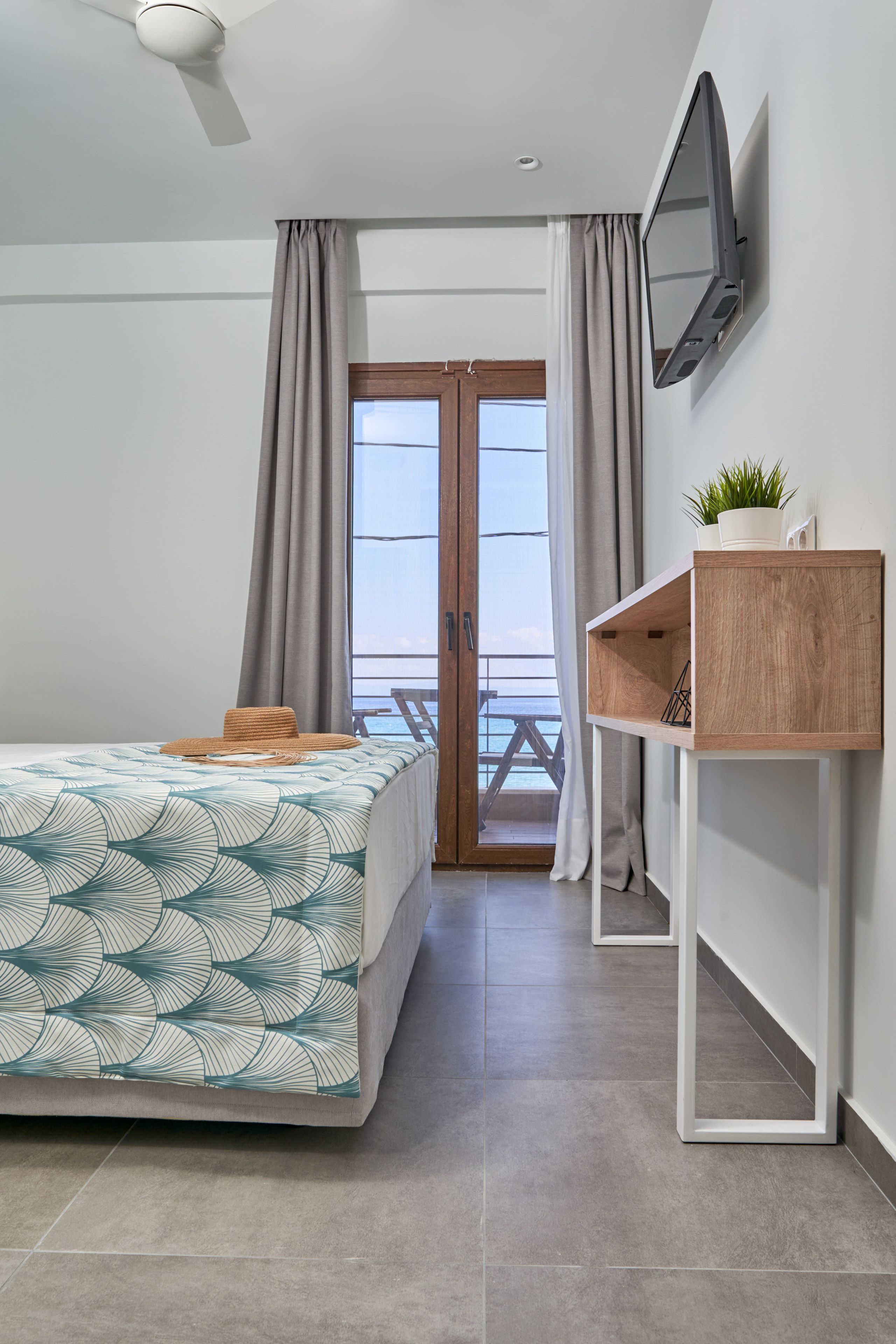 A modern hotel room with a bed, a TV mounted on the wall, a wooden side table with a plant, and a balcony with a view.