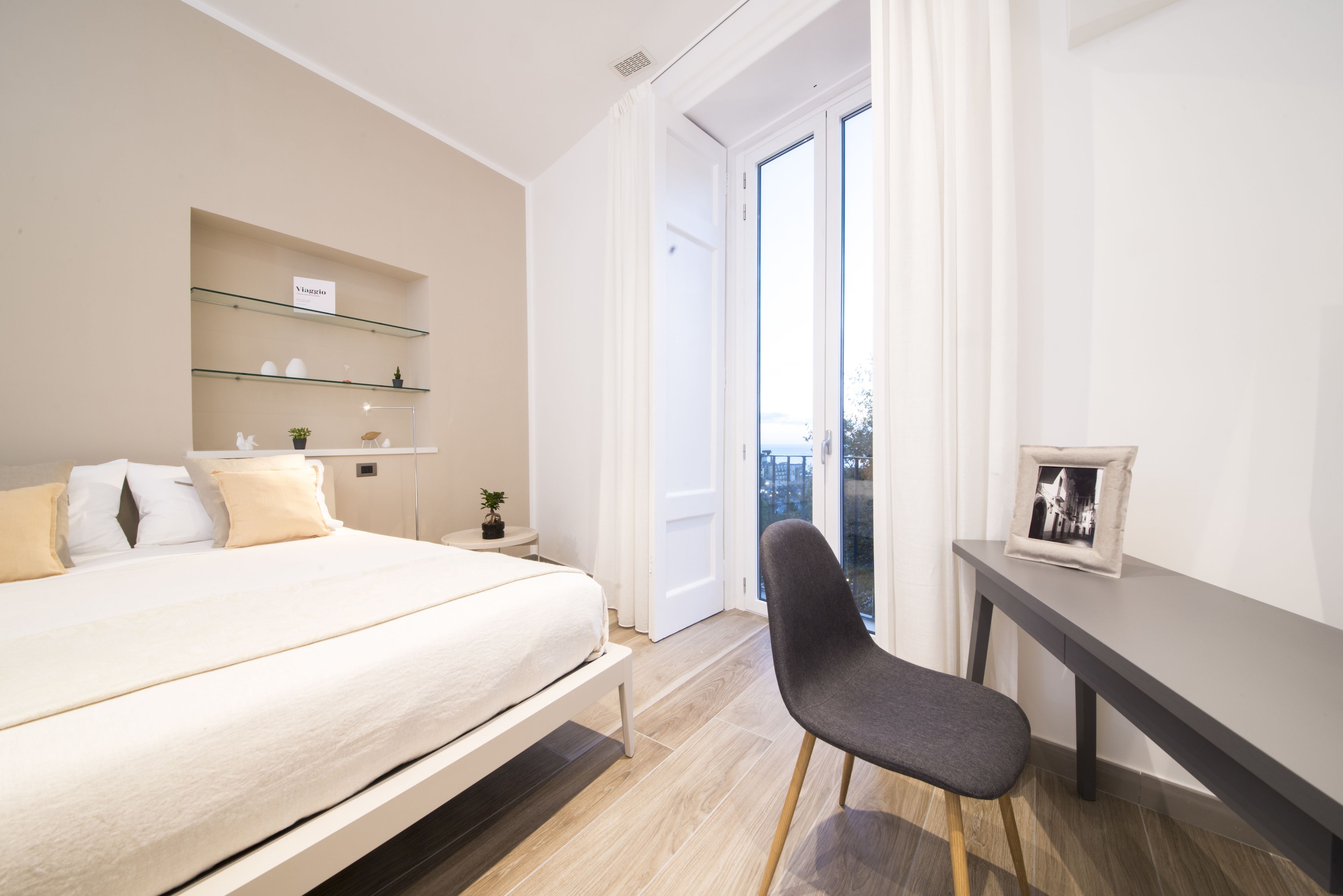 Ein modernes Hotelzimmer mit Bett, Nachttischen, Schreibtisch und Stuhl. Ein Fenster mit Vorhängen bietet Blick auf die Stadt.